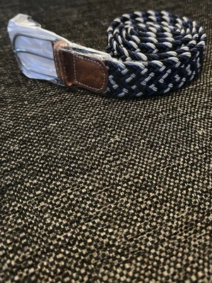 Flätat marinblått bälte med läderdetalj - Snyggt flätat bälte i marinblått, vitt och grått med silverfärgat spänne och bruna läderdetaljer vid spännet. Bältet är elastiskt och passar perfekt till jeans eller chinos för en avslappnad men stilren look.