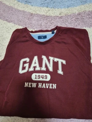 Vinröd sweatshirt från GANT - Snygg vinröd sweatshirt från GANT med tryck 'GANT 1949 NEW HAVEN' på bröstet. Klassisk rund halsringning och avslappnad passform. Perfekt för en casual look och enkel att matcha med jeans eller joggers.
