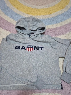 Gant hoodie - Snygg gråmelerad hoodie från Gant med klassisk logga och sköld framtill. Tröjan har huva, ribbade muddar och är tillverkad i mjukt material som känns skönt mot huden. Perfekt för en avslappnad och sportig stil.