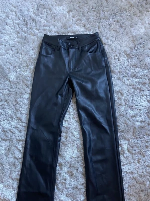 Svarta skinnbyxor från BikBok - Snygga svarta skinnbyxor från BikBok i rak modell. Byxorna har klassisk femficksdesign, knapp och dragkedja framtill samt bälteshällor. Perfekta för dig som vill ha en edgy look med stilren känsla.