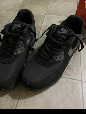 Svarta Nike Air Max sneakers - Snygga svarta Nike Air Max sneakers med grå detaljer och klassisk swoosh på sidan. Skorna har snörning, rund tå och platt sula. Tillverkade i en mix av syntet och textil för en sportig look. Perfekta för dig som gillar streetwear och vill ha bekväma sneakers.