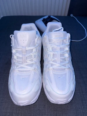 Nike vita sneakers med snörning - Fräscha helvita Nike sneakers med klassisk snörning och meshpaneler för extra andningsförmåga. Snygg sportig design med rund tå och diskret Nike-logga på sidan. Perfekta för dig som gillar stilrena och cleana skor. Pris kan diskuteras