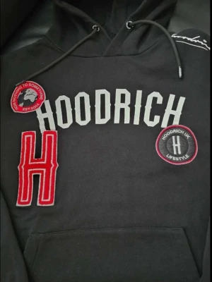 Svart hoodie från Hoodrich - Svart hoodie med stora tryck och broderade patches framtill. HOODRICH-logga i vitt, röd broderad H och två runda patches med text och logga. Klassisk känguruficka och dragsko i huvan. Perfekt för streetwear-stil. Den är använd 1 gång vid intresse kan ni alltid ställa frågor. OM NI VILL HA FLER BILDER SKRIV I DM! 