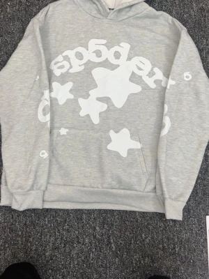 Grå hoodie med stjärnor från Sp5der - Snygg grå hoodie från Sp5der med stora vita stjärnor och logga tryckt över hela framsidan. Hoodien har en klassisk huva och magficka, samt ribbade muddar vid ärmar och nederkant. Perfekt för en avslappnad och trendig streetwear-look.