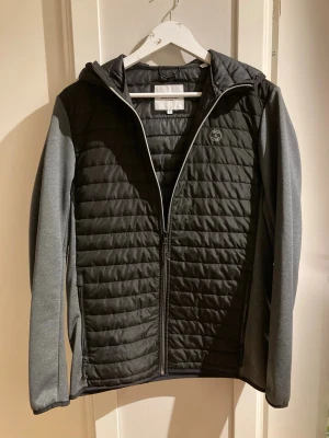 Svart vindjacka från Jack & Jones - Snygg svart vindjacka från Jack & Jones med quiltad front och gråa ärmar. Jackan har huva, dragkedja framtill och diskret logga på bröstet. Perfekt för dig som vill ha en stilren och funktionell jacka till vardags.