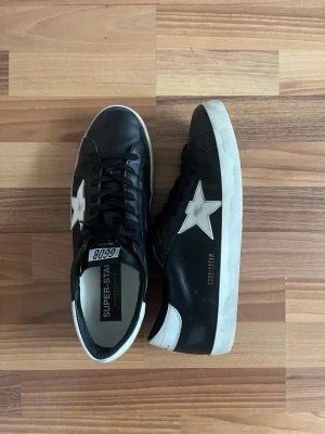 Svarta Golden Goose skor - Feta Golden Goose skor i svart skinn med vit stjärndetalj på sidan och vit häl. Klassisk låg modell med snörning och platt sula. Perfekta för en cool och avslappnad stil. Storlek 45. Inte använda mycket! Fråga gärna!