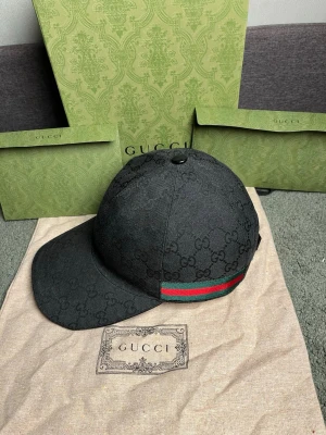 Gucci svart keps med GG-monogram - Svart keps från Gucci med klassiskt GG-monogram över hela kepsen. På sidan finns en ikonisk grön och röd rand. Kepsen har böjd skärm och är tillverkad i canvasmaterial. Perfekt för dig som vill ha en stilren accessoar med lyxig känsla.