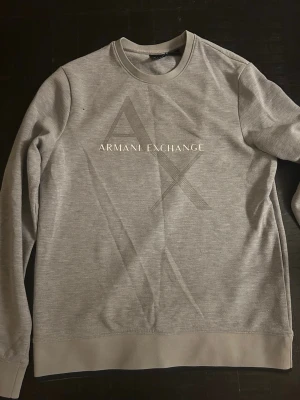 Grå sweatshirt från Armani Exchange - Snygg grå sweatshirt från Armani Exchange med stort AX-tryck och logga på bröstet. Tröjan har rund halsringning, långa ärmar och ribbade muddar vid ärmslut och nederkant. Perfekt för en clean och stilren look.