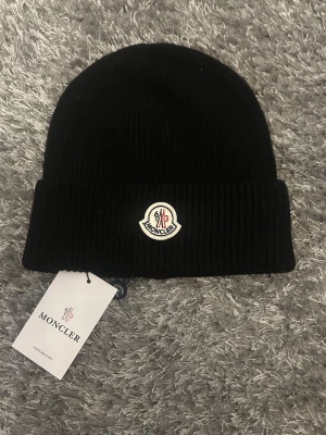 Svart Moncler mössa i ribbstickad ull - Säljer en svart ribbstickad mössa från Moncler med klassisk uppvikt kant och Moncler-logga framtill. Mössan är tillverkad i mjuk ull och har en stilren, enkel design som passar perfekt för kalla dagar.