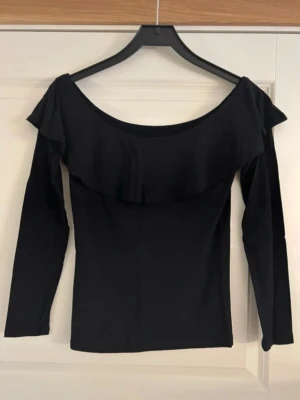 Svart offshoulder topp med volang - Säljer en svart offshoulder topp med lång ärm och bred volang längs halsringningen. Inga defekter och jättefin på. 