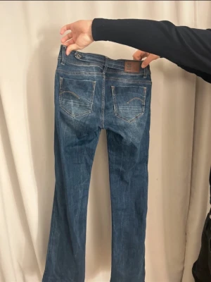 G-Star RAW Midge Bootcut jeans blå - Säljer ett par G-Star RAW Midge Bootcut jeans i klassisk blå tvätt. Modellen har bootcut-ben, fem fickor och snygga detaljer på bakfickorna. Jeansen är tillverkade i slitstarkt denim och har normal passform. Perfekt för dig som gillar en tidlös look.
