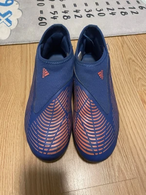 Blå-orange adidas fotbollsskor - Säljer ett par blå fotbollsskor från adidas med orangea detaljer och mönster. Skorna har en strömlinjeformad design utan snörning, med mönstrade linjer på ovansidan och tre klassiska ränder på sidan. Materialet är syntet och textil, perfekt för gräsplan. Man kan också diskutera priset 