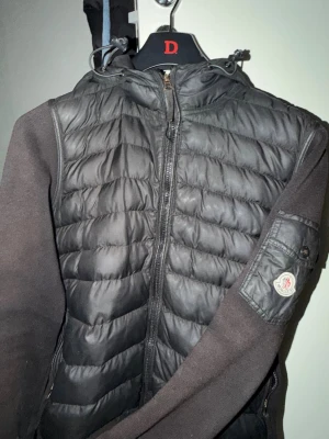 Svart Moncler pufferjacka med huva - Svart pufferjacka från Moncler med quiltad front och huva. Jackan har dragkedja hela vägen, ribbade muddar och Moncler-logga på ärmen. Materialmix med polyester och stickade partier på ärmarna ger en schysst kontrast. Perfekt för dig som gillar street och vill ha något snyggt till hösten.