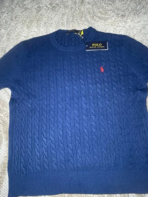 Blå stickad tröja från Polo Ralph Lauren - Säljer en marinblå stickad tröja från Polo Ralph Lauren med klassiskt kabelmönster och röd broderad logga på bröstet. Rund halsringning och ribbade muddar vid ärmslut och nederkant. Perfekt för dig som gillar stilrena och tidlösa plagg.