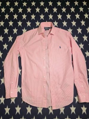 Rosa Raffe skjorta - Stilig, rosa-vit randig skjorta från Ralph Lauren i strl S. Nyskick förutom en liten skråma på höger arm som knappast syns, förutom vid noga inspektion. Skriv om du har någon fundering!