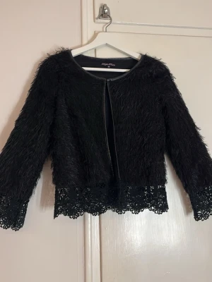 Svart pälsjacka med spetsdetaljer - Säljer en svart pälsjacka från Boohoo med fluffig yta och snygga spetsdetaljer vid ärmslut och nederkant. Jackan är långärmad och har en öppen front utan knappar eller dragkedja. Perfekt för dig som vill sticka ut med en unik look.