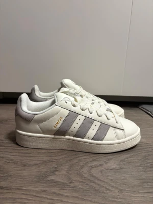 Adidas Campus sneakers i vitt och grått 00s - Säljer ett par Adidas Campus sneakers i vitt med ljusgrå detaljer och klassiska tre ränder på sidan. Skorna har snörning, platt sula och rund tå. Materialet är en mix av skinn och mocka, med Adidas-logga på tungan och hälen. Perfekt för dig som gillar stilrena och tidlösa sneakers. Skorna är helt oanvända, precis ute ur boxen. Skorna kom i fel storlek och ångerrätt var över tiden. 