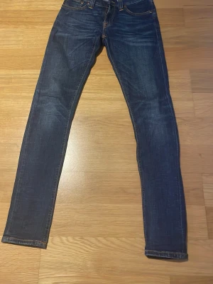 Nudie Jeans Tight Long John 25/32 - Mörkblå skinny jeans från Nudie Jeans, modell Tight Long John. Tillverkade i 98% ekologisk bomull och 2% elastan för en stretchig och bekväm passform. Klassisk femficksdesign med orangea kontrastsömmar och låg midja.