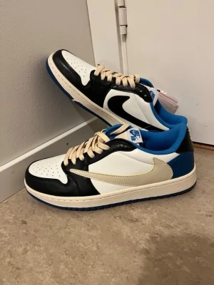 Nike Jordan 1 Low Travis Scott blå/svart - Nike Jordan 1 Low Travis Scott med svart, vit och blå färg. Skorna har beige snörning, omvänd Swoosh i vitt och klassisk Jordan 1 siluett. Yttermaterialet är skinn och insidan är fodrad med blå textil. Perfekt för dig som gillar sneakers med unik design.