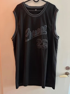 Chicago basketlinne XXXL svart/grå - Snyggt basketlinne i svart med grå detaljer och texten 'Chicago' på bröstet samt Michael Jordans siffra 23 både fram och bak. Linningen har grå och svarta ränder och materialet är mesh som andas. Perfekt för dig som gillar sportig stil och vill sticka ut.