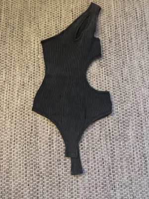 Svart oneshoulder ribbad bodysuit - Säljer en svart ribbad bodysuit med oneshoulder-design och cool cut-out detalj på sidan. Materialet är mjukt och stretchigt. Passformen är tight och toppen är ärmlös med asymmetrisk form.
