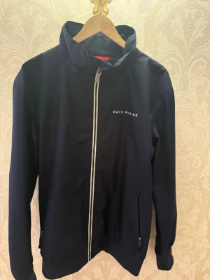 Mörkblå vindjacka Race Marine L. Ej använd  - Snygg mörkblå vindjacka från Race Marine med vit dragkedja och diskret logga på bröstet. Jackan har hög krage, långa ärmar och två fickor med dragkedja. Perfekt för blåsiga dagar och enkel att matcha med jeans eller sneakers.