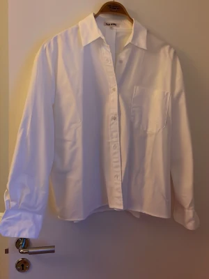 Vit skjorta från Djerf Avenue (S) - Vit skjorta från Djerf Avenue i storlek S. Klassisk modell med krage, lång ärm och bröstficka med broderad logga. Skjortan har knappar framtill och är gjord i mjuk bomull som känns skön mot huden. Perfekt för dig som gillar stilrena och enkla plagg.