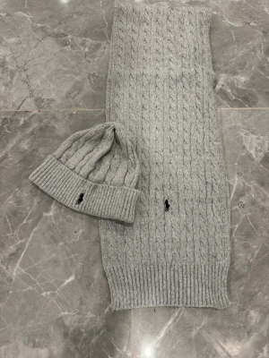 Grå stickad väst från Polo Ralph Lauren - Säljer en grå stickad väst från Polo Ralph Lauren med klassiskt kabelstickat mönster och svart broderad logga på framsidan. Västen har ribbad nederkant och är gjord i mjukt ullmaterial som känns skönt mot huden. Perfekt för lager-på-lager under kyliga dagar.