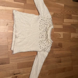 Vit fluffig tröja med paljetter - Säljer en vit fluffig långärmad tröja med glittriga paljetter över bröstet och axlarna. Tröjan har rund hals och är gjord i ett mjukt, pälsliknande material som känns riktigt cozy. Perfekt för dig som gillar lite extra bling i vardagen!