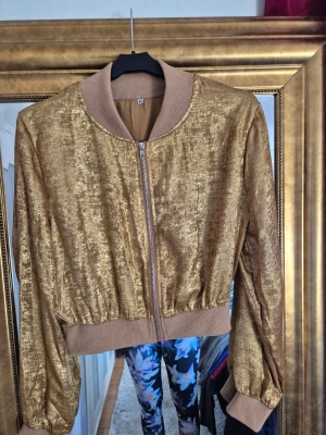 Guldig bomberjacka, M - Säljer en unik bomberjacka i guldig nyans med ribbade muddar och krage. Jackan har dragkedja framtill och en glansig, lyxig finish som verkligen sticker ut. Perfekt för dig som vill ha något extra i garderoben och gillar att synas.