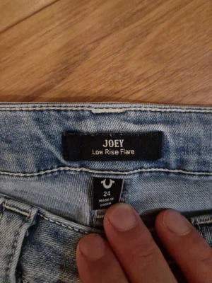 True Religion Joey Low Rise Flare Jeans - Säljer ett par ljusblå jeans från True Religion, modell Joey Low Rise Flare. De har låg midja och utsvängda ben för en snygg retrostil. Klassisk femficksdesign och slitningar som ger en cool vibe. Perfekta för dig som gillar statement-jeans.