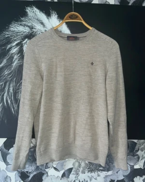 Beige merinoulltröja från Morris - Snygg beige stickad tröja från Morris i 100% merinoull. Klassisk rund hals och diskret logga broderad på bröstet. Perfekt för dig som gillar stilrena och mjuka plagg med lång ärm och ribbade muddar.