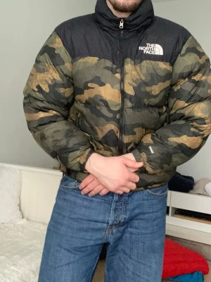 Camouflage pufferjacka från The North Face - Säljer en fet pufferjacka från The North Face i storlek M. Jackan har camouflage-mönster i grönt, brunt och svart med svart axelparti och tydlig logga på bröstet och ryggen. Hög krage och dragkedja framtill. Perfekt för kalla dagar.