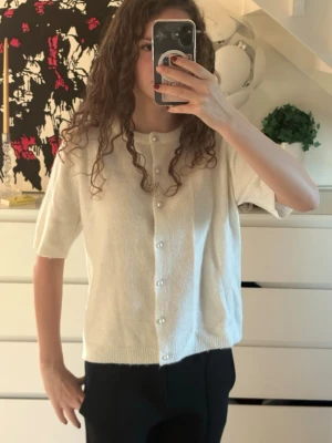 Vit kofta med pärlknappar - Superfin vit kofta med kort ärm ifrån Lager 157 i storlek S. Koftan har pärlknappar hela vägen framtill och är stickad i ett mjukt material. Perfekt att slänga på sig över ett linne eller t-shirt för en chill look.