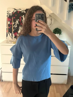 Blå tröja med volangärmar - Säljer en blå tröja med volangdetaljer på ärmarna. Tröjan har rund halsringning och trekvartsärm. Den är i storlek 164 men är som en Xs. Perfekt för dig som vill ha något stilrent men ändå med en twist.