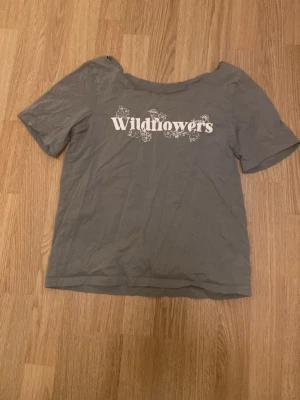 Grå t-shirt med Wildflowers-tryck - Säljer en grå t-shirt med vit text 'Wildflowers' och blommönster framtill. T-shirten har korta ärmar och en avslappnad passform. Perfekt för dig som gillar enkel men snygg stil. Materialet känns som bomull och är mjukt mot huden.