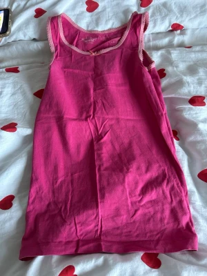 Rosa linnetopp från Pepperts - Säljer en rosa linnetopp från Pepperts i storlek 134/140. Toppen har ljusrosa spetskant runt halsringningen och axelbanden samt en liten rosett framtill. Materialet är mjuk bomull med lite elastan för extra komfort. Perfekt för varma dagar!