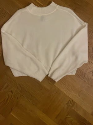 Vit stickad tröja med hög krage - Säljer en kort vit stickad tröja med hög ribbad krage och breda muddar vid ärmslut och nederkant. Tröjan har en loose passform och är långärmad, perfekt för lager-på-lager under kyliga dagar. Enkel och stilren design som funkar till många outfits. Sträcker sig precis ner till naveln ungefär.