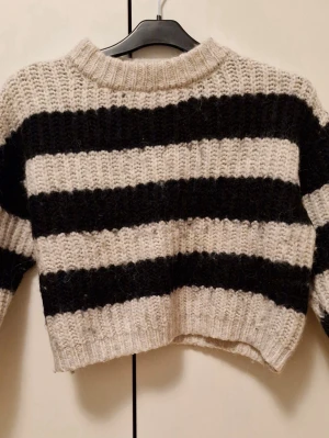 (Barnkläder) Randig croppad stickad tröja Zara - Säljer en croppad stickad tröja från Zara med breda svarta och beige ränder. Storleken är 140 cm. Den är väldigt nopprig. Tröjan har rund hals och lång ärm, perfekt för dig som gillar en chill och trendig stil. Grovstickad och mjuk, passar till jeans eller kjol.