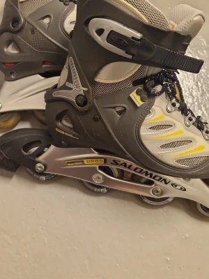 Salomon inlines med skydd - Säljer ett par grå och svarta Salomon inlines med gula detaljer, robust snörning och spänne för extra stöd. Medföljer ett set silverfärgade skydd i meshväska, perfekt för att rulla runt i parken eller på gatan. Skorna har ventilerande mesh och slitstark plast för komfort och säkerhet. Pris kan såklart diskuteras!