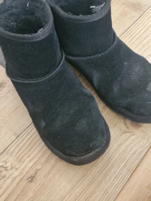 Svarta mockaboots med rund tå - Mysiga svarta boots i mocka med rund tå och platt sula. Skorna har en enkel design utan snörning och är fodrade för extra värme. Perfekta för kalla dagar och passar till många olika outfits. Det finns smuts som kan tvättas bort och märken där bakom. Annars går det att använda