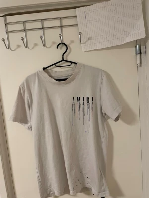 Vit AMIRI t-shirt med tryck - Säljer en vit t-shirt från AMIRI med coolt drip-tryck både fram och bak. T-shirten har korta ärmar och en klassisk rund hals. Materialet är mjuk bomull och trycket har färgstänk i blått, svart och orange för en unik vibe. Liknande modell som original. 
