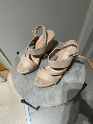 Beige kilklack sandaler från Gogo Shoes - Snygga beige sandaler med kilklack från Gogo Shoes. Skorna har breda korslagda remmar och öppen tå, samt justerbart spänne vid hälen. Yttermaterialet är mocka och innersulan är mjuk för extra komfort. Perfekt för dig som gillar stilrena och somriga vibes.