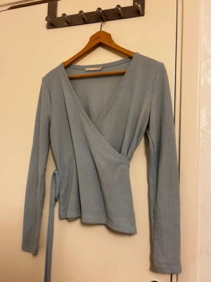 Ljusblå omlottopp från 157 - Säljer en ljusblå omlottopp från 157 i ribbad bomull. Toppen har långa ärmar, v-ringad hals och knytband i midjan för en snygg passform. Hålet på höger sidan är för att träa snöret igenom och knyta vid midjan. Perfekt att styla med jeans eller kjol för en chill look.