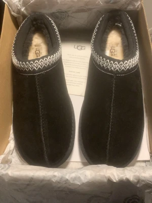 Svarta UGG slip-on skor med mönster - Säljer ett par svarta slip-on skor från UGG med mjuk päls på insidan och snyggt broderat mönster i vitt runt öppningen. Skorna har en rund tå och platt sula med tydligt mönster undertill. Utsidan är i mocka och insidan är fodrad med päls för extra komfort.