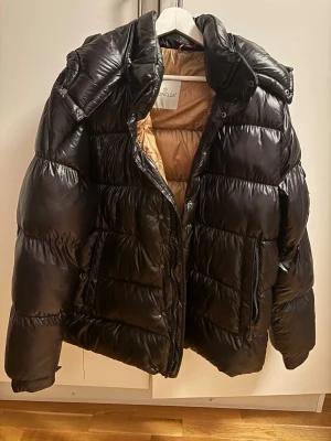 Svart Moncler pufferjacka med beige foder - Säljer en svart pufferjacka från Moncler med glansig finish och beige/guld foder. Perfekt skick. Sparsamt använd. Storlek 4. Jackan har huva, dragkedja och tryckknappar framtill. Mycket varm, perfekt för kalla dagar och har en riktigt schysst oversized vibe.