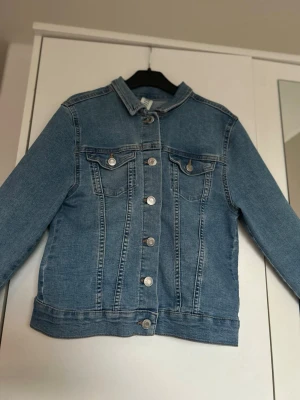  blå jeansjacka från H&M - Säljer en blå jeansjacka från H&M storlek 152. Det är raka linjer och silverfärgade knappar. Jackan har två bröstfickor med  knapp. Perfekt att slänga på sig över en hoodie eller t-shirt. Kontakta om ni är intresserad