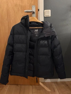 Svart pufferjacka från Helly Hansen - Säljer en svart pufferjacka från Helly Hansen med stor huva och diskret HH-logga på bröstet och ryggen. Jackan är vadderad och har dragkedja framtill. Perfekt för kalla dagar och riktigt skön att bära. Passformen är normal och jackan är långärmad. (Liten svart fläck från tuschpenna på insidan)