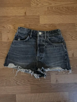 Grå denimshorts från Zara - Säljer ett par gråa denimshorts från Zara i storlek 32. Modellen är högmidjad med klassiska fem fickor och råa, fransiga kanter nedtill. Shortsen har knapp och dragkedja framtill samt bälteshällor. Perfekta för en chill look under varma dagar.