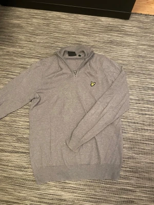 Grå stickad half zip från Lyle & Scott - Säljer en grå stickad tröja från Lyle & Scott i storlek M. Tröjan har half zip, långärmad design och klassisk gul fågel-logga på bröstet. Perfekt för dig som gillar stilrena och bekväma plagg med snygga detaljer.